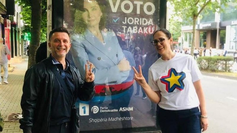 Erion Braçe dhe Delina Ibrahimaj pozojnë me dy gishtat lartë para posterit elektoral të Jorida Tabakut 
